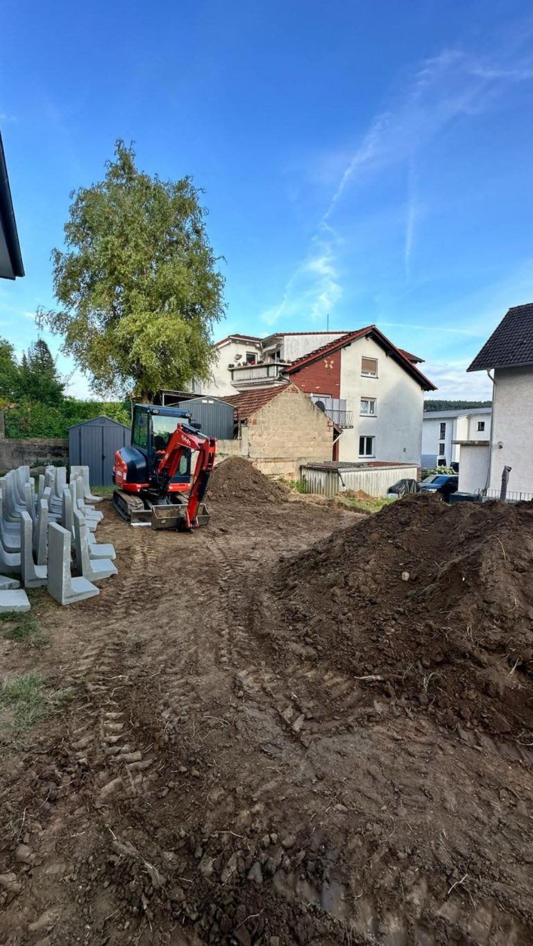 Bauplatz mit Bagger, Erde und Häusern im Hintergrund. Klare blaue Himmel.