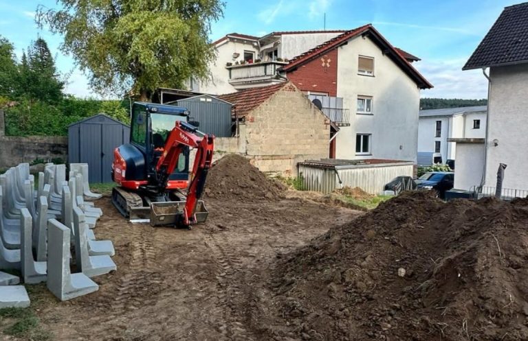 Bagger und Erdhaufen auf Baustelle hinter Häusern.