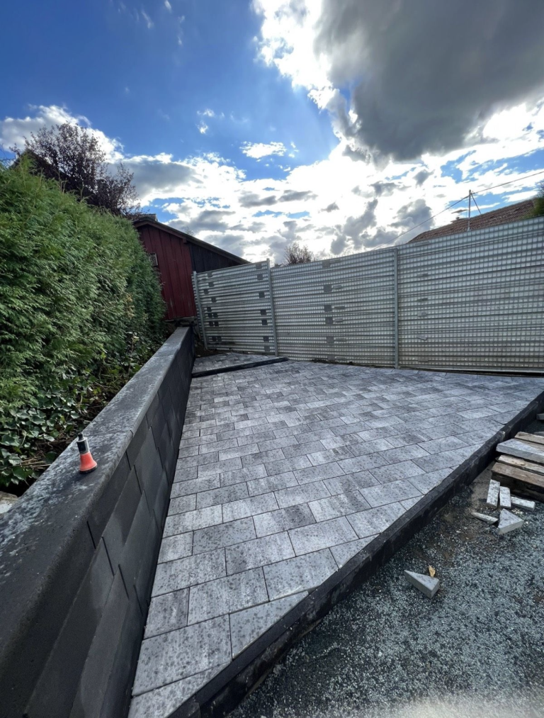 Gepflasterter Bereich mit grauen Steinplatten und Mauer im Hintergrund, blauer Himmel.