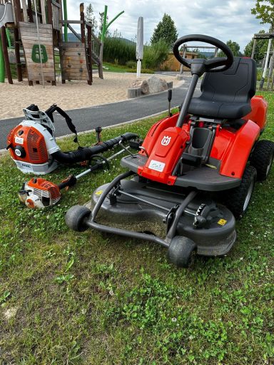 Rasenmäher und Motorsense auf einer Wiese neben einem Spielplatz.