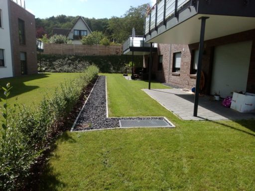 Grüne Rasenfläche mit Pflanzbeet und moderner Wohngebäude-Architektur im Hintergrund.