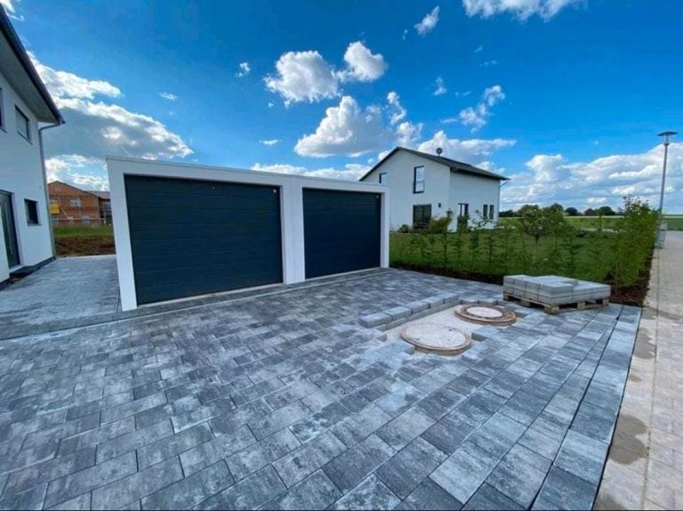 Zwei Garagen mit dunkelblauen Toren, umgeben von einem gepflasterten Hof und Pflanzen.